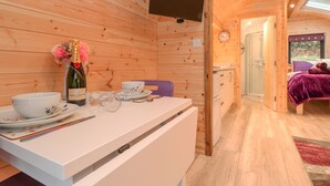 A wooden cabin interior with a dining area, a TV mounted on the wall, and a visible bathroom with a shower. - Lovies Place - Crossgate Luxury Glamping (Penrith)