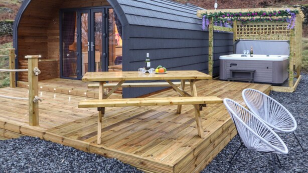 A cozy wooden pod structure with a picnic table and chairs, a hot tub, and a garden area with flowers. - Lovies Place - Crossgate Luxury Glamping (Penrith)