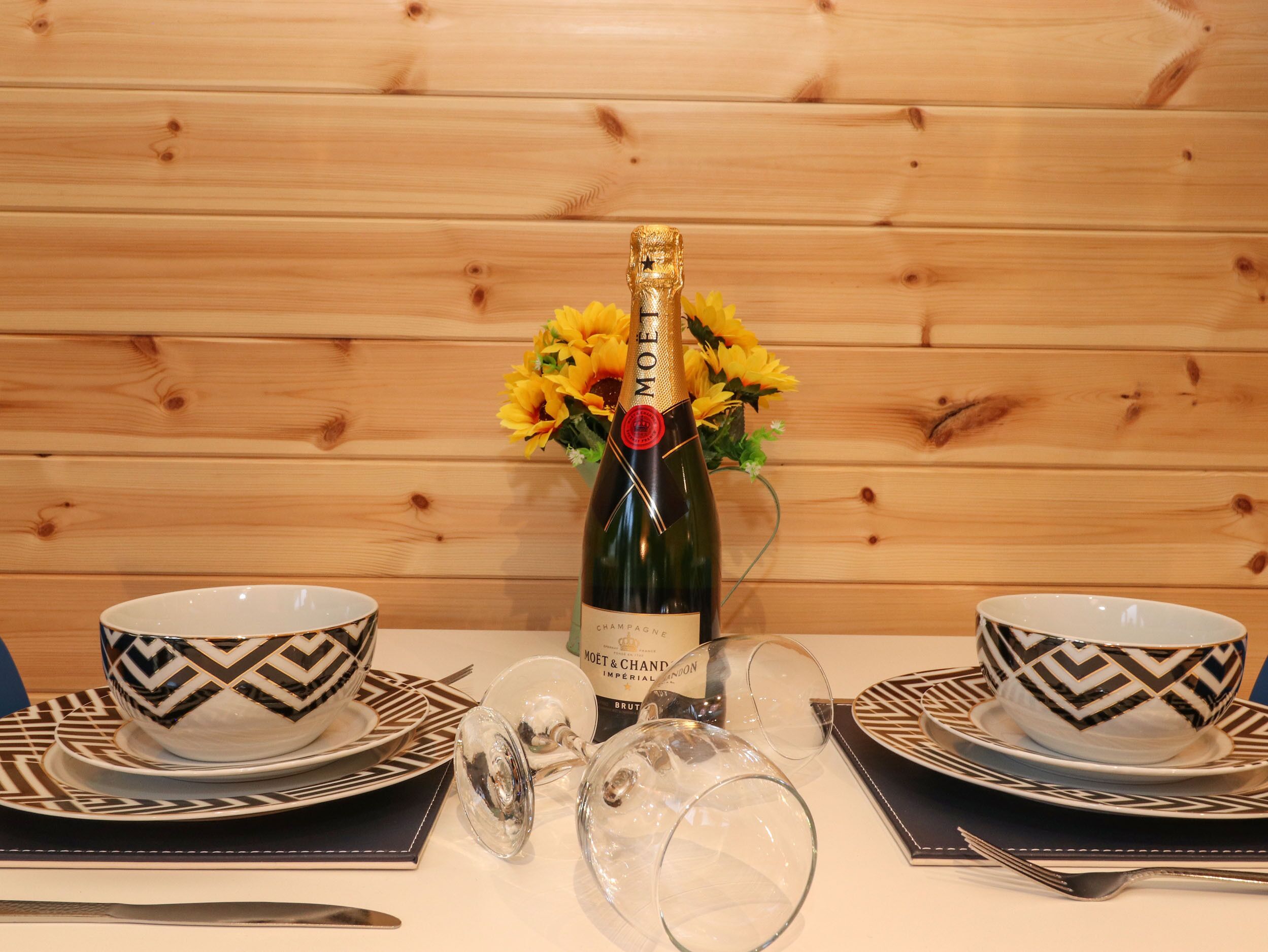 Een gedekte tafel met een champagnefles, twee decoratieve schalen en een vaas vol gele bloemen.