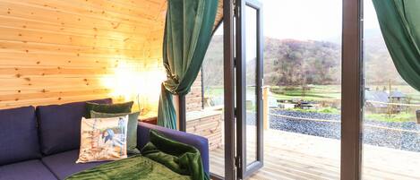 A cozy room with a wooden ceiling, a blue sofa, a green throw blanket, and a view of a rural landscape through a glass door.