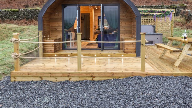A wooden cabin with a glass door entrance, a deck with rope barrier, and a hot tub in the background.