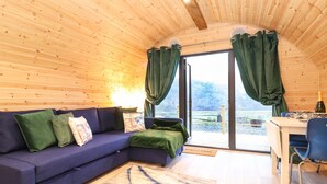 A cozy cabin interior with a wooden ceiling, a blue sofa, a dining table with chairs, and a view through a glass door.