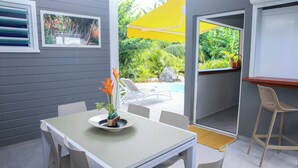 A patio with a table and chairs, a yellow umbrella, and a view of a pool and greenery.
