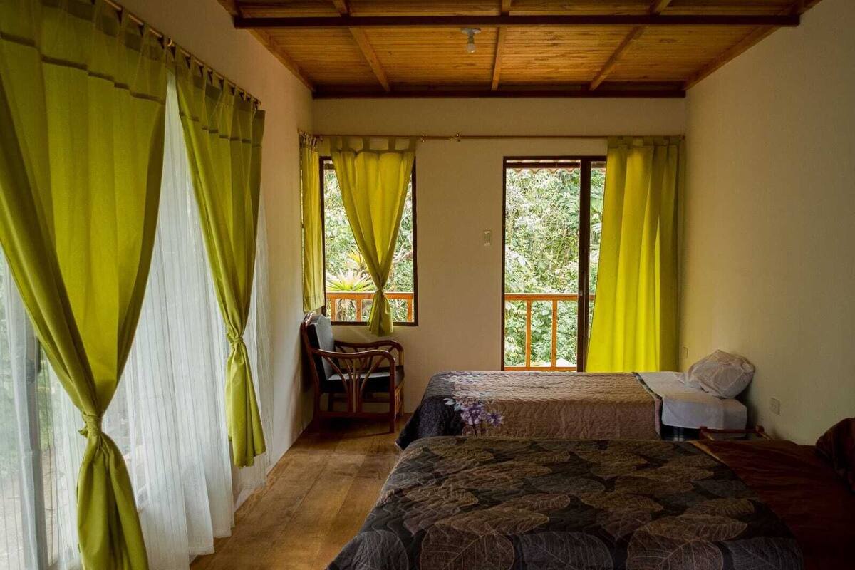A bedroom with two beds, a wooden ceiling, and large windows with curtains.