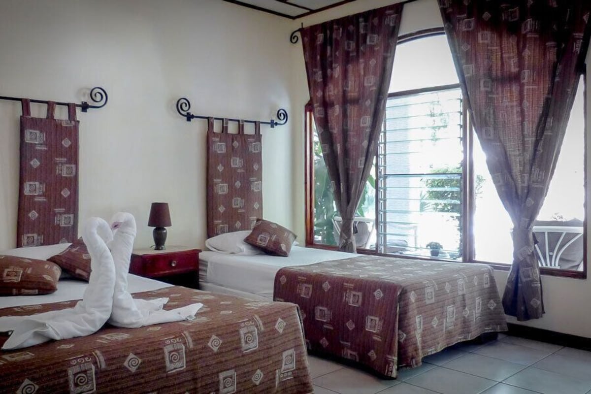 A hotel room with two beds, a nightstand, a lamp, and a window with curtains.