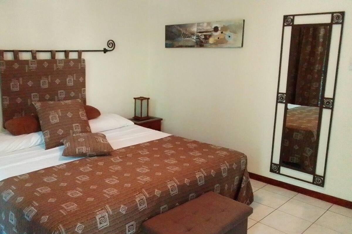 Un dormitorio con cama, mesita de noche, lámpara, espejo y un cuadro en la pared.