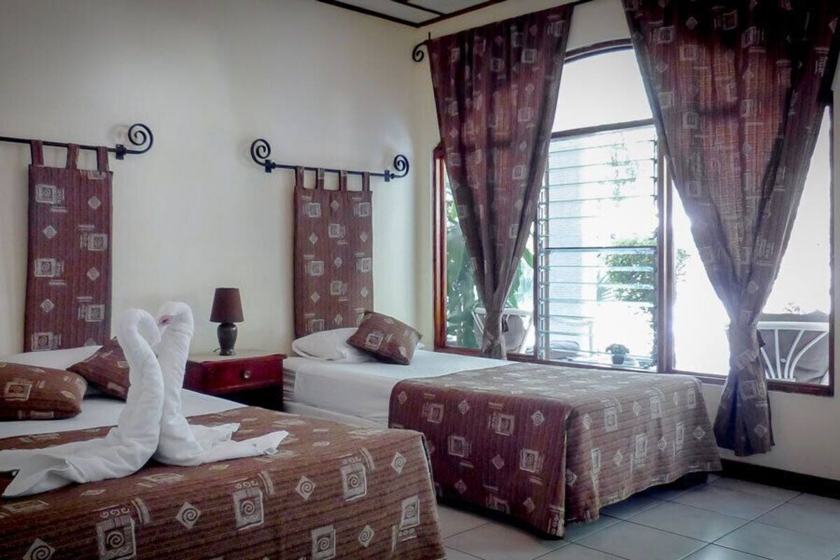 Una habitación de hotel con dos camas, una mesita de noche, una lámpara y una ventana con cortinas.