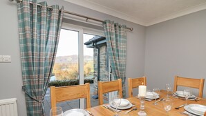 Uma sala de jantar com mesa posta para quatro pessoas, cadeiras de madeira, uma vela e vista para uma paisagem montanhosa através de uma janela com cortinas xadrez.