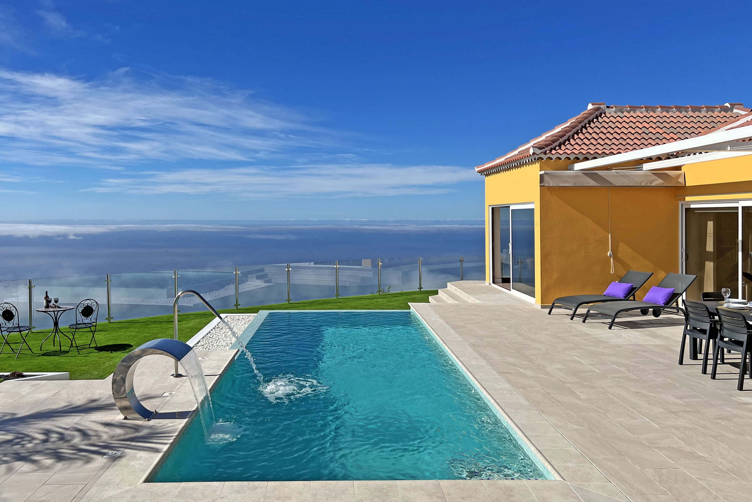 A swimming pool with a clear view of the horizon, a patio area with chairs and a table, and a building with a tiled roof.