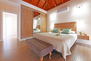 Un dormitorio con techo de madera, una cama grande con colcha blanca y beige, una mesita de noche de madera y un armario de madera con puerta corredera.