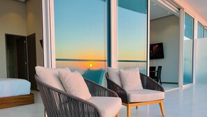A modern room with a bed, two wicker chairs with cushions, a TV, and a glass wall with a sunset view. - Beachfront 3BR-3BA SunsetParadise (Flamingos)