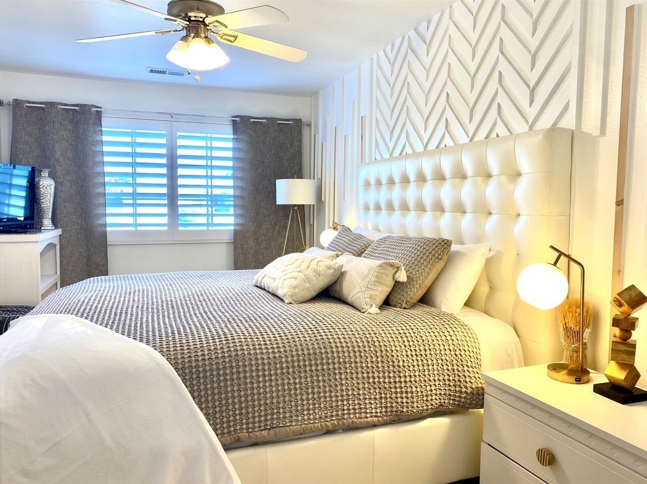 A bedroom with a tufted headboard, a bed with patterned bedding, a bedside lamp, and a nightstand with a lamp.
