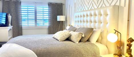 A bedroom with a tufted headboard, a bed with patterned bedding, a bedside lamp, and a nightstand with a lamp.
