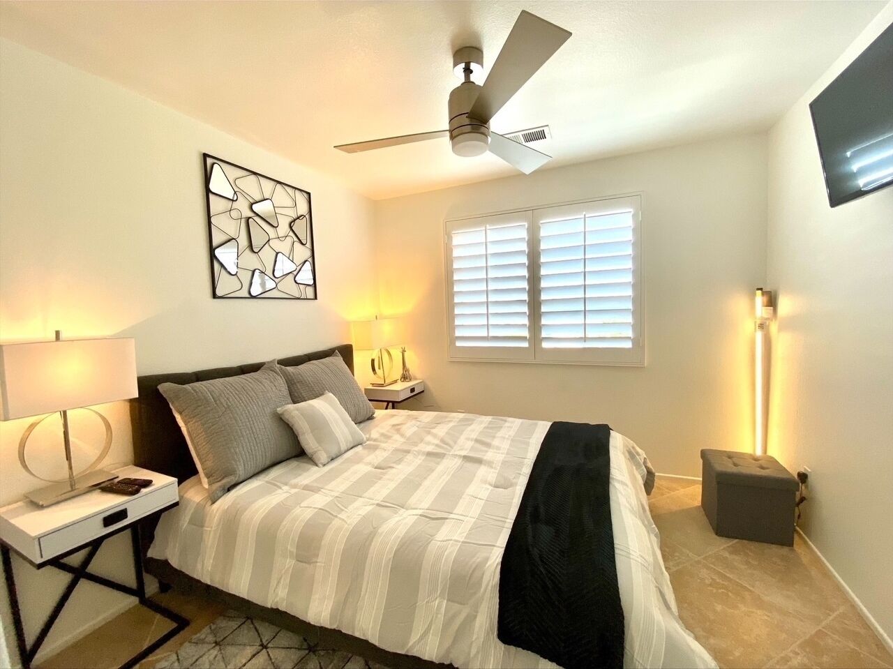 A bedroom with a bed, bedside lamps, a ceiling fan, a window with blinds, and a wall-mounted TV.