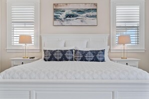 Une chambre avec un lit blanc, deux tables de chevet, un tableau représentant des vagues et des stores blancs.
