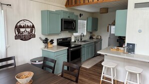 A modern kitchen with teal cabinets, a black microwave, and a dining area with a wooden table and chairs. - Lakeside cottage, water access + pet friendly with additional fee (Jasper)
