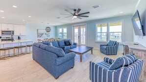 Een ruime woonkamer met een grote blauwe bank, gestreept fauteuils, een salontafel en een eettafel. Er is een keuken met witte kasten en een magnetron.