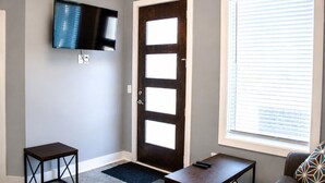 Ein Zimmer mit einer dunkelholzigen Tür, einem an der Wand befestigten Flachbildfernseher, einem kleinen Tisch und einem Fenster mit Jalousien.