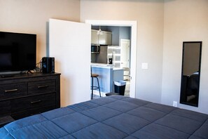 Quarto com uma cama, cômoda, televisão e uma kitchenette ao fundo.