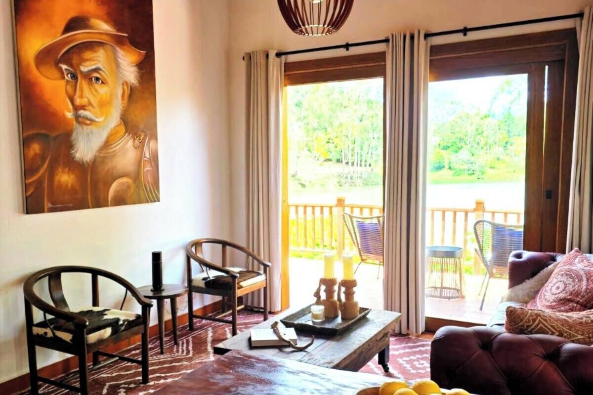 A cozy living room with a wooden ceiling, a large painting on the wall, a dining table with chairs, and a view of the outdoors through a glass door.