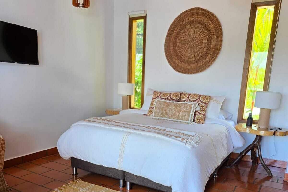 A bedroom with a bed, a woven wall hanging, two windows, and a lamp on a bedside table.