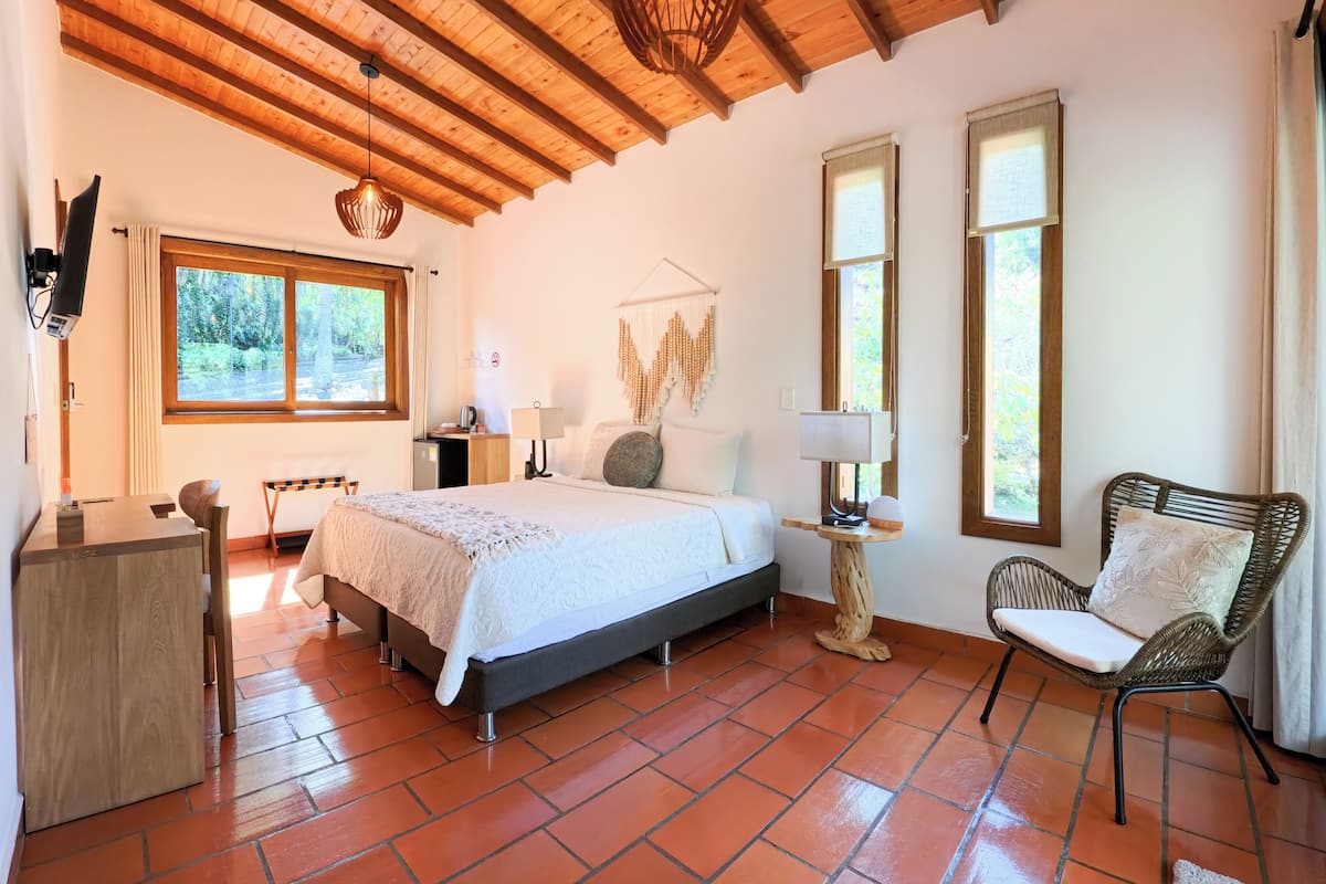 A bedroom with a bed, bedside table, lamp, chair, and a view of the outdoors through a window.