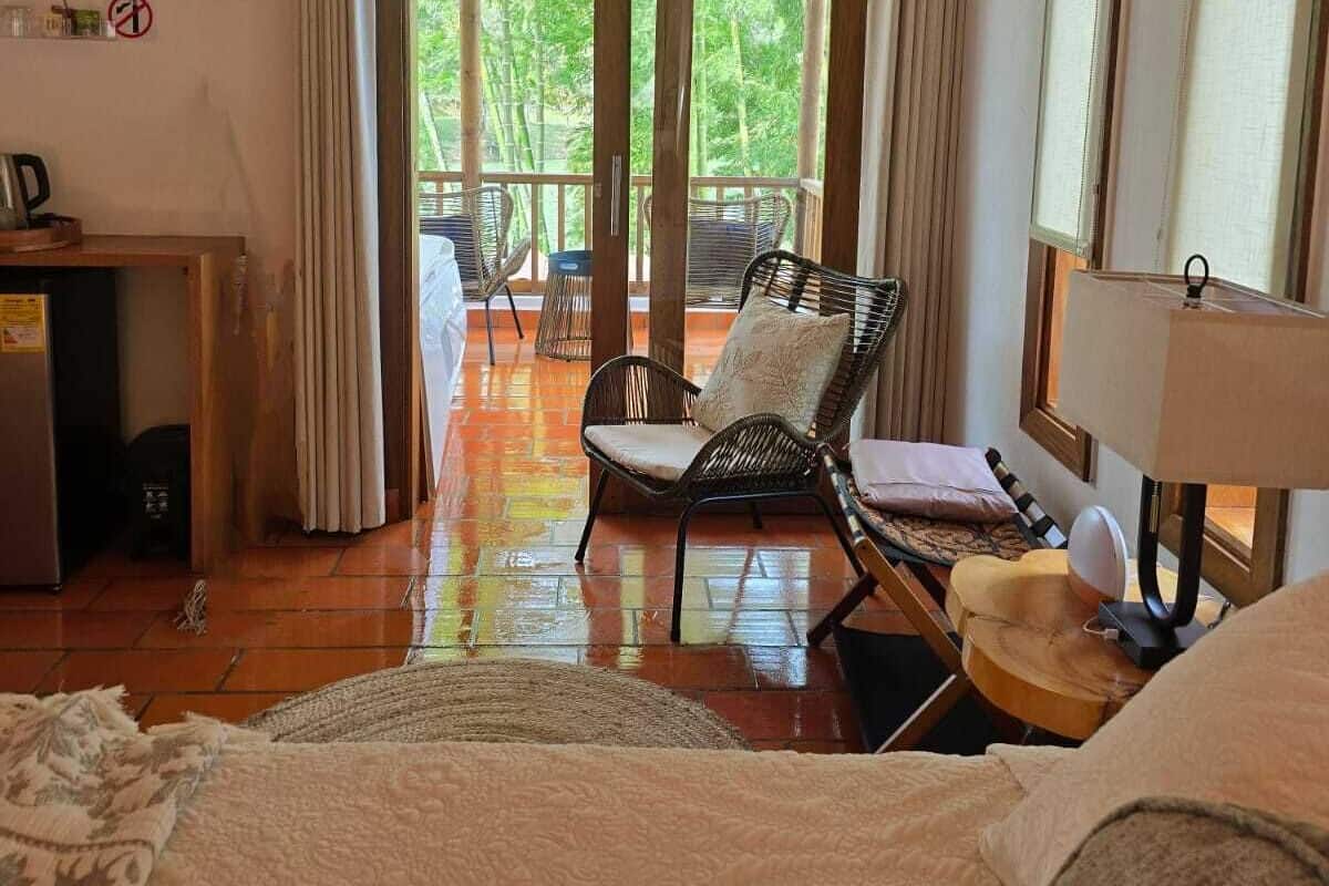 A room with a bed, a chair, a table, a lamp, and a view of trees through a glass door.