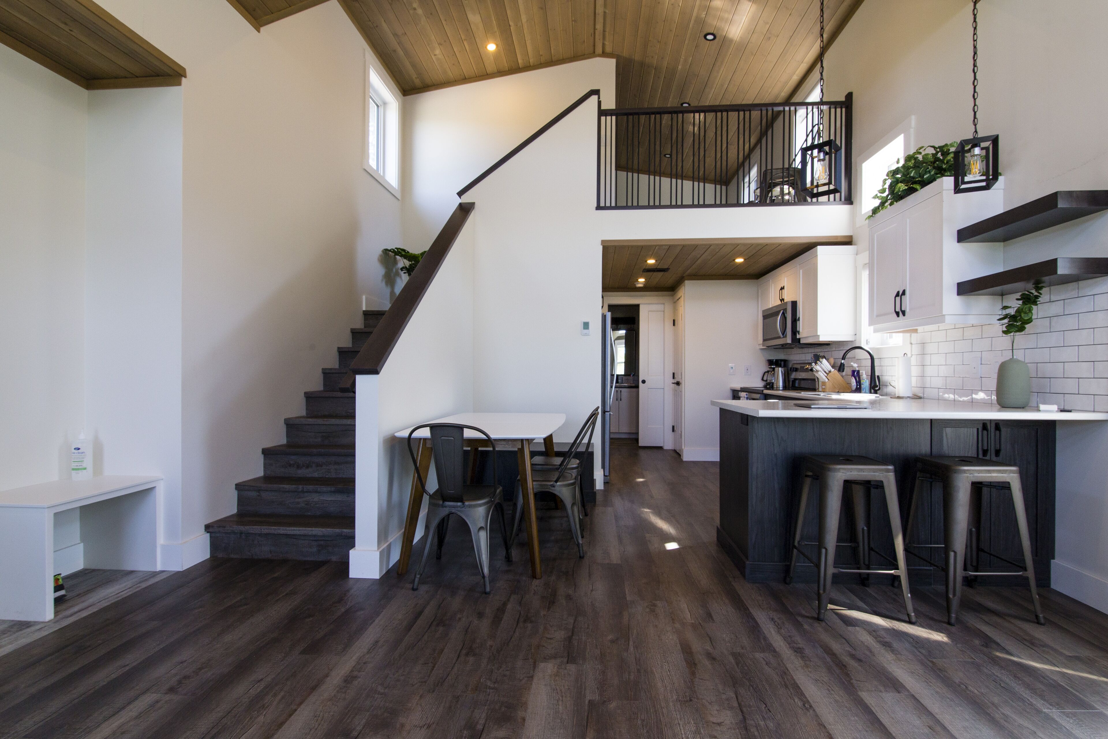 A modern kitchen with a dark wood floor, a central island with stools, and a staircase leading to an upper level.