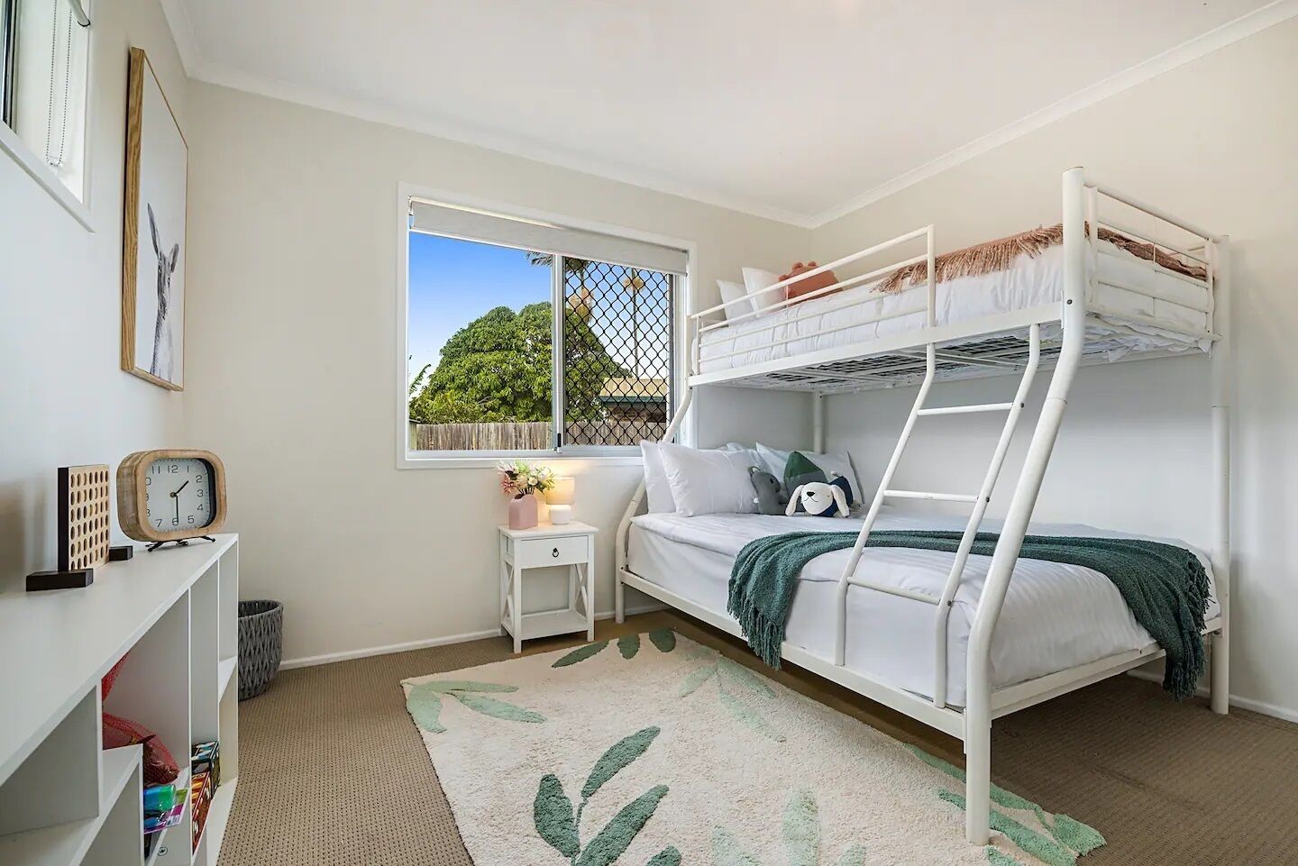 A bunk bed room with a window, a small bedside table, a clock, and a bookshelf.
