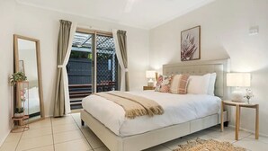A bedroom with a large bed, a nightstand, a bedside table, a mirror, and a view of the outdoors.