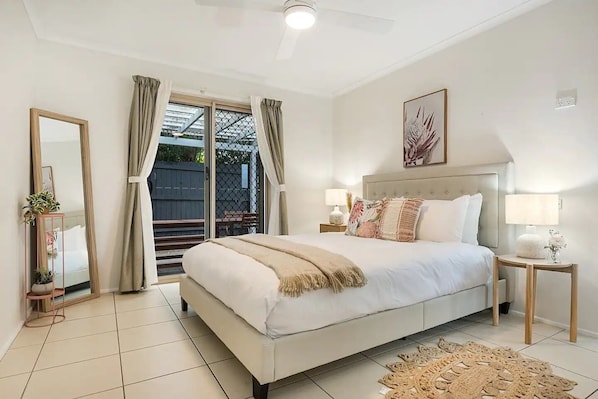 A bedroom with a large bed, a nightstand, a bedside table, a mirror, and a view of the outdoors.