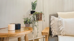 A cozy living room with a round wooden table, a grey sofa, and a white couch. There are three potted plants on a shelf and a woven basket on the table.