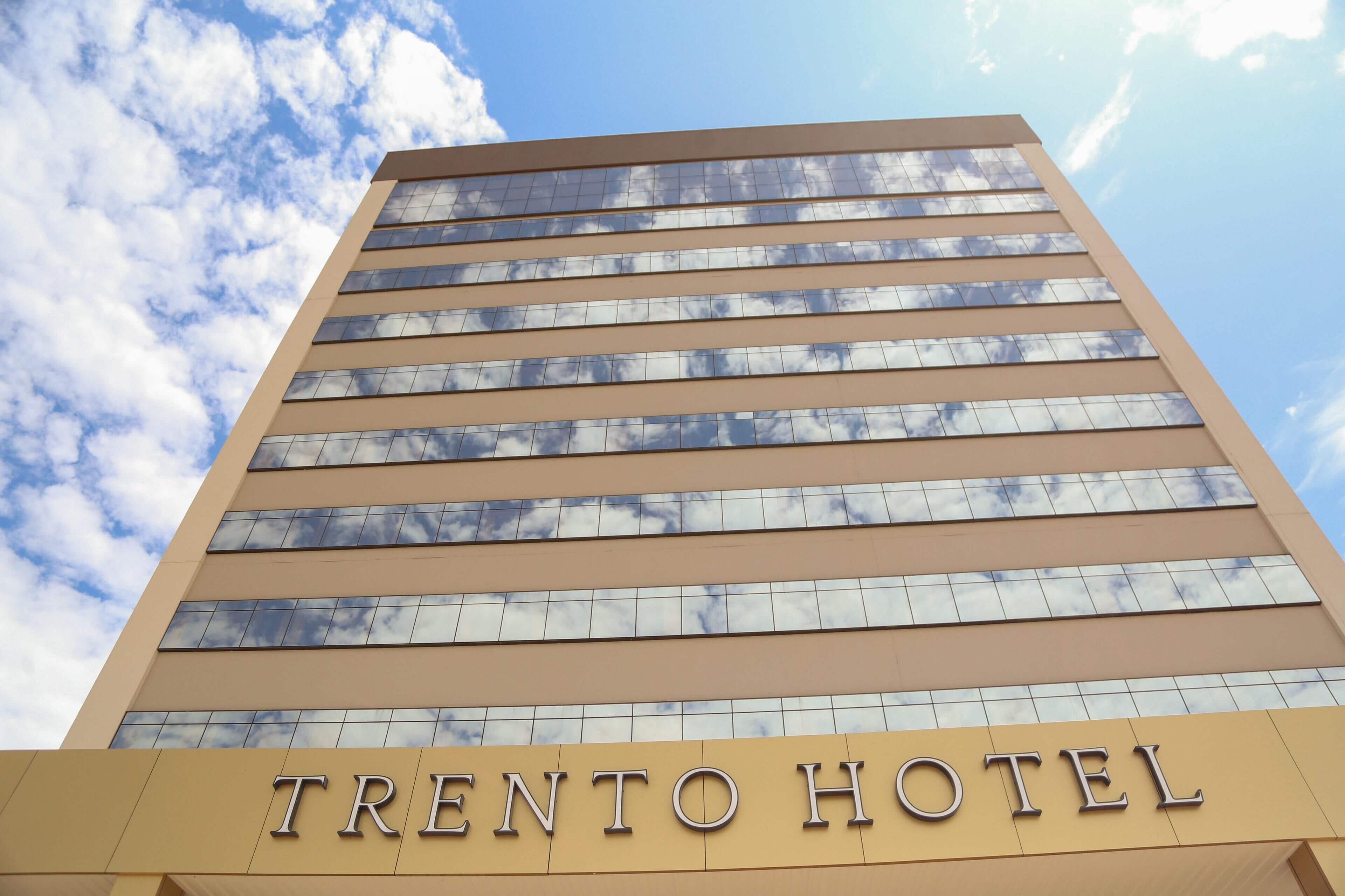 The exterior of the Trento Hotel with a clear sky and clouds reflected in the windows.