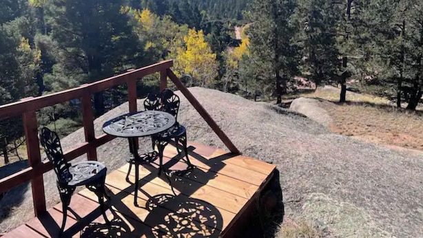 Um deck de madeira com mesa e cadeiras, com vista para uma paisagem arborizada.