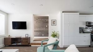 A modern living room with a TV, wooden flooring, a teal armchair, and a kitchen area with white cabinets and appliances. - `Bike` at Hills Meaford | 1BD Suite + Pets (Meaford)