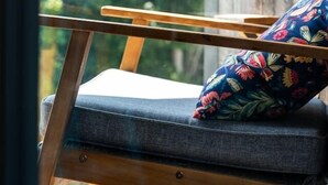 Una silla de madera con un cojín y un almohadón floral, ubicada cerca de una ventana con vista a follaje.