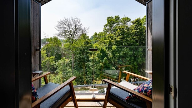 Un balcón con dos sillas de madera y una mesa, con vistas a un bosque verde y un cielo despejado.