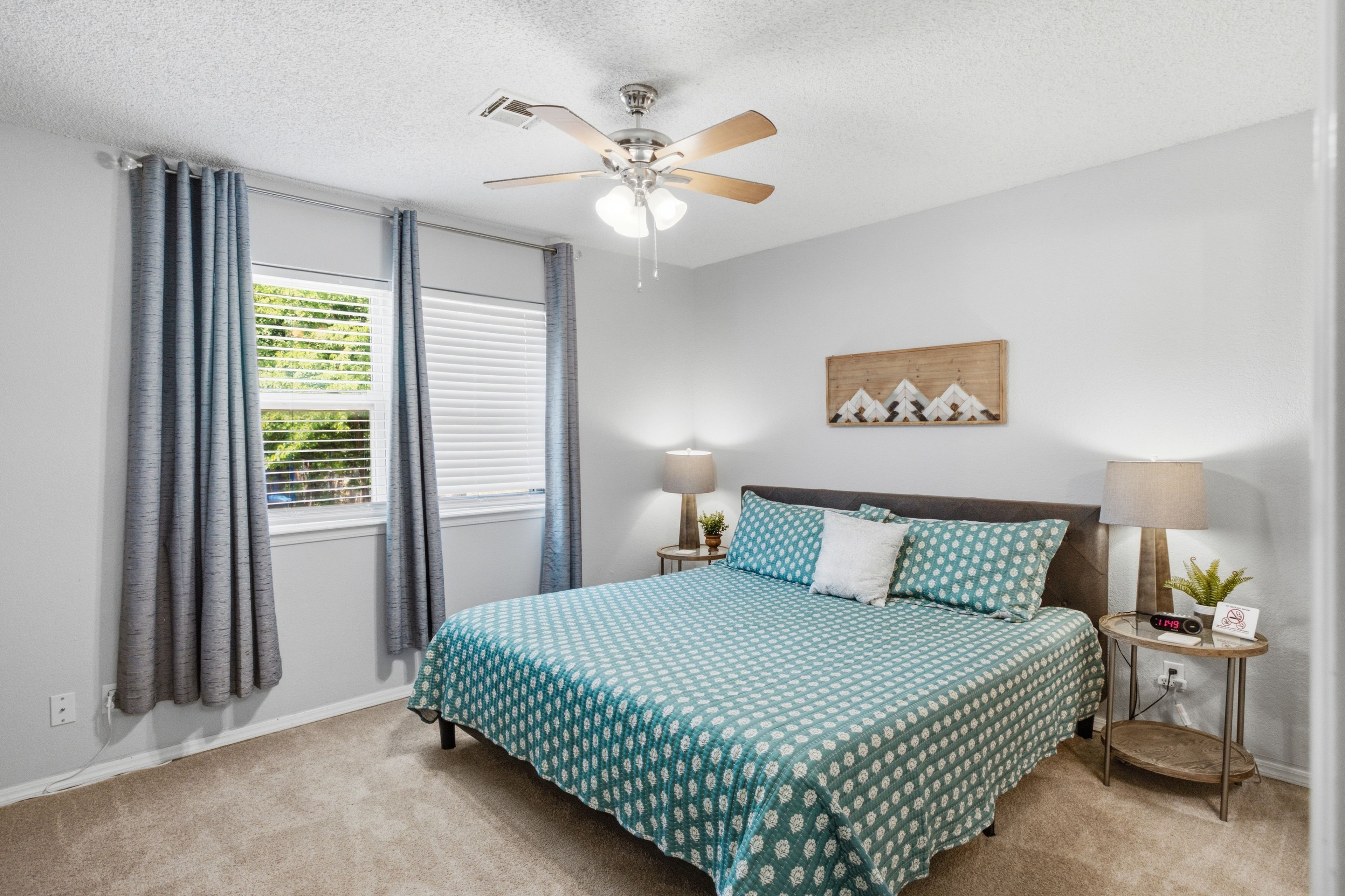 A bedroom with a bed, a ceiling fan, a window with curtains, a bedside table with a lamp, and a wall-mounted artwork.