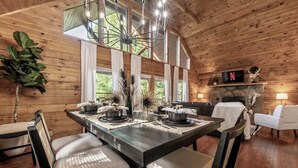 A cozy wooden interior with a dining table set for four, surrounded by chairs. A fireplace is visible in the background, and a television is mounted above it. - Sip your coffee in the rocking chairs while enjoying the serene Mountain View’s. (Sevierville)