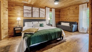 A wooden-paneled bedroom with a bed, a sofa, and a nightstand. - Sip your coffee in the rocking chairs while enjoying the serene Mountain View’s. (Sevierville)