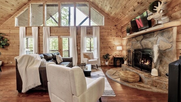 A cozy cabin interior with a stone fireplace, a leather sofa, and a television. - Sip your coffee in the rocking chairs while enjoying the serene Mountain View’s. (Sevierville)