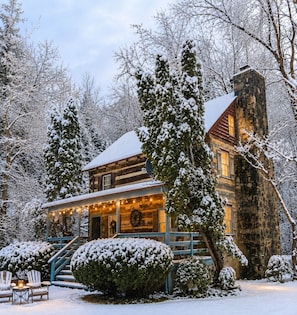 A log cabin with a stone chimney, surrounded by snow-covered trees and a porch with string lights.