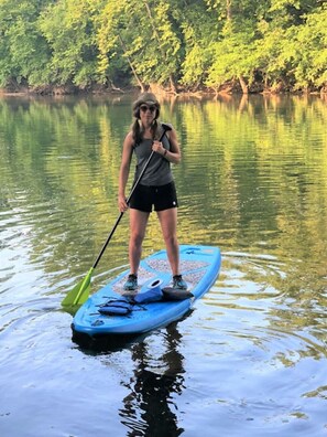 Una persona de pie en una tabla de paddleboard sobre aguas tranquilas, con árboles al fondo.