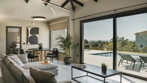 A modern living room with a sofa, coffee table, and dining area. There is a large window with a view of a pool and greenery. - Single-Storey Contemporary Villa, A/C, Private Pool and Panoramic Sea Views - 10 mins to Beaches (Avithos)