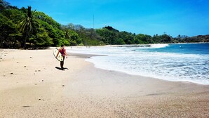 Una playa con una persona cargando una tabla de surf, rodeada de abundante vegetación y aguas azules cristalinas.