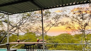 Un espacio de estar techado al aire libre con una mesa y sillas, con vistas a un paisaje pintoresco con árboles y un atardecer.