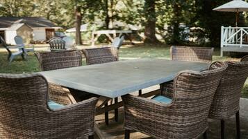 A rectangular outdoor table with wicker chairs, surrounded by trees and a white fence.