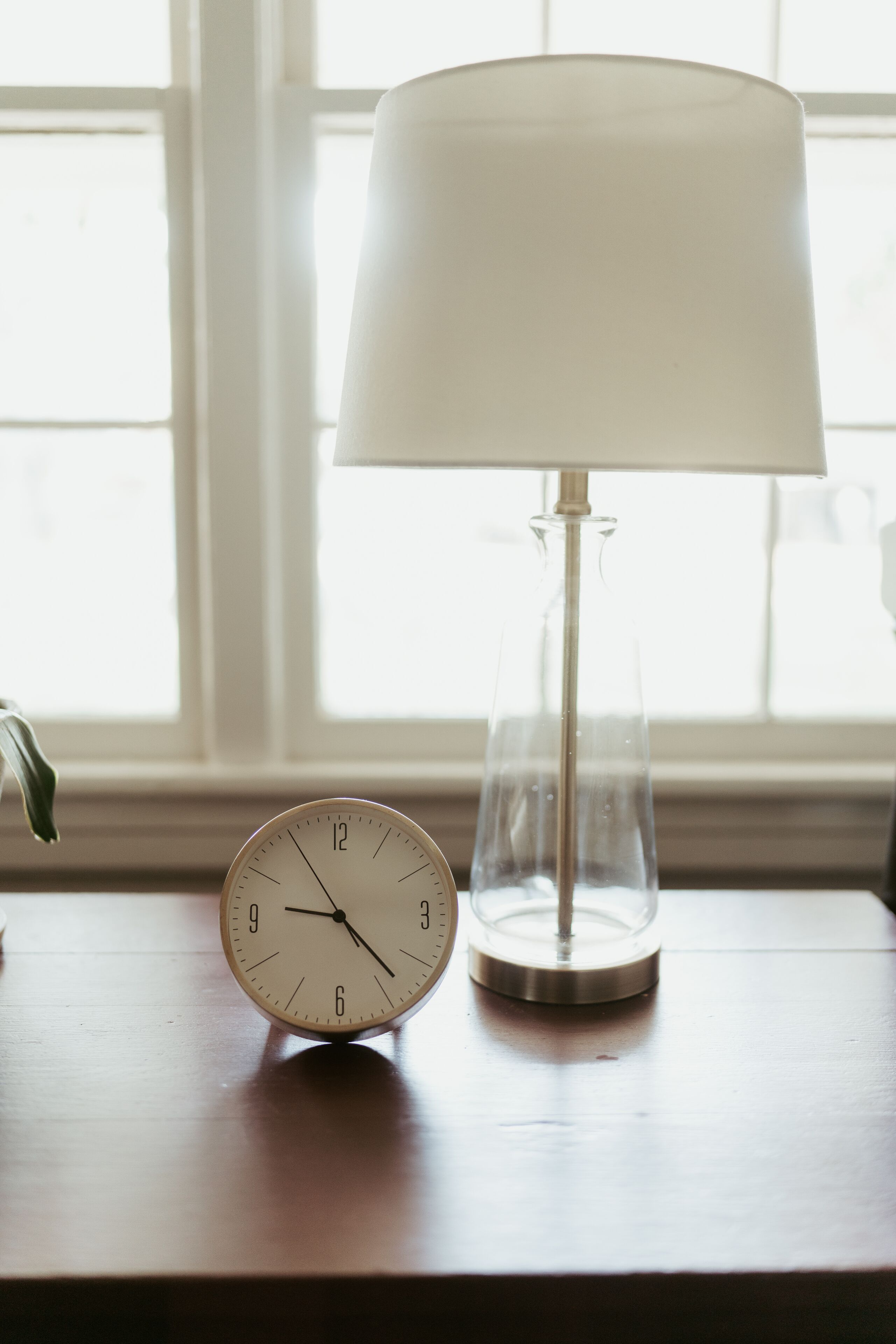 Un reloj y una lámpara sobre una mesa de madera cerca de una ventana.
