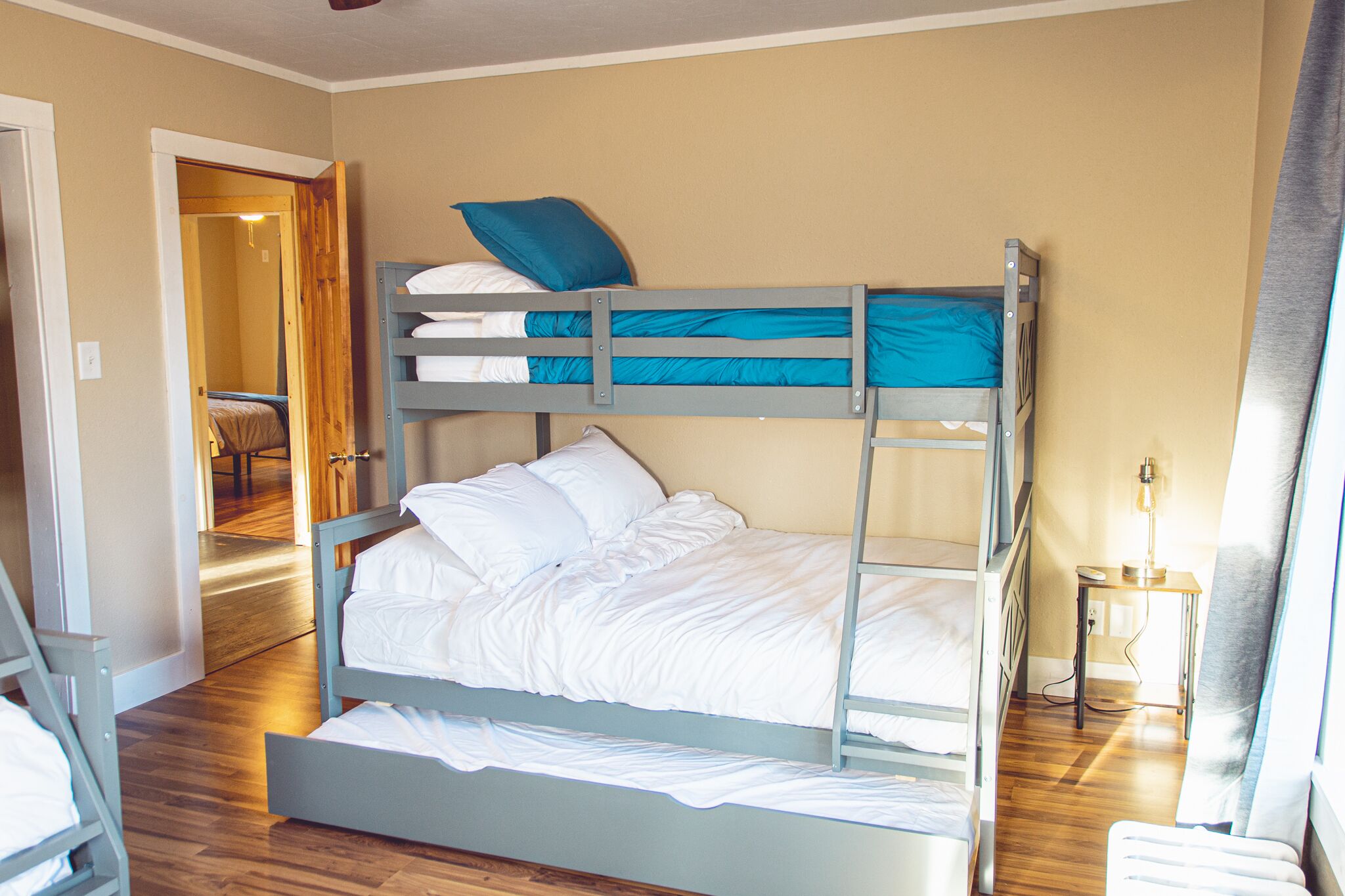 Een slaapkamer met stapelbedden, een houten vloer, een bureau met lamp en een deur naar een andere kamer.