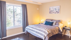 A bedroom with a bed, a bedside table with a lamp, a window with curtains, and a painting on the wall. - Lakeside Luxury on Upper & Lower Post Lake Channel (Post Lake)